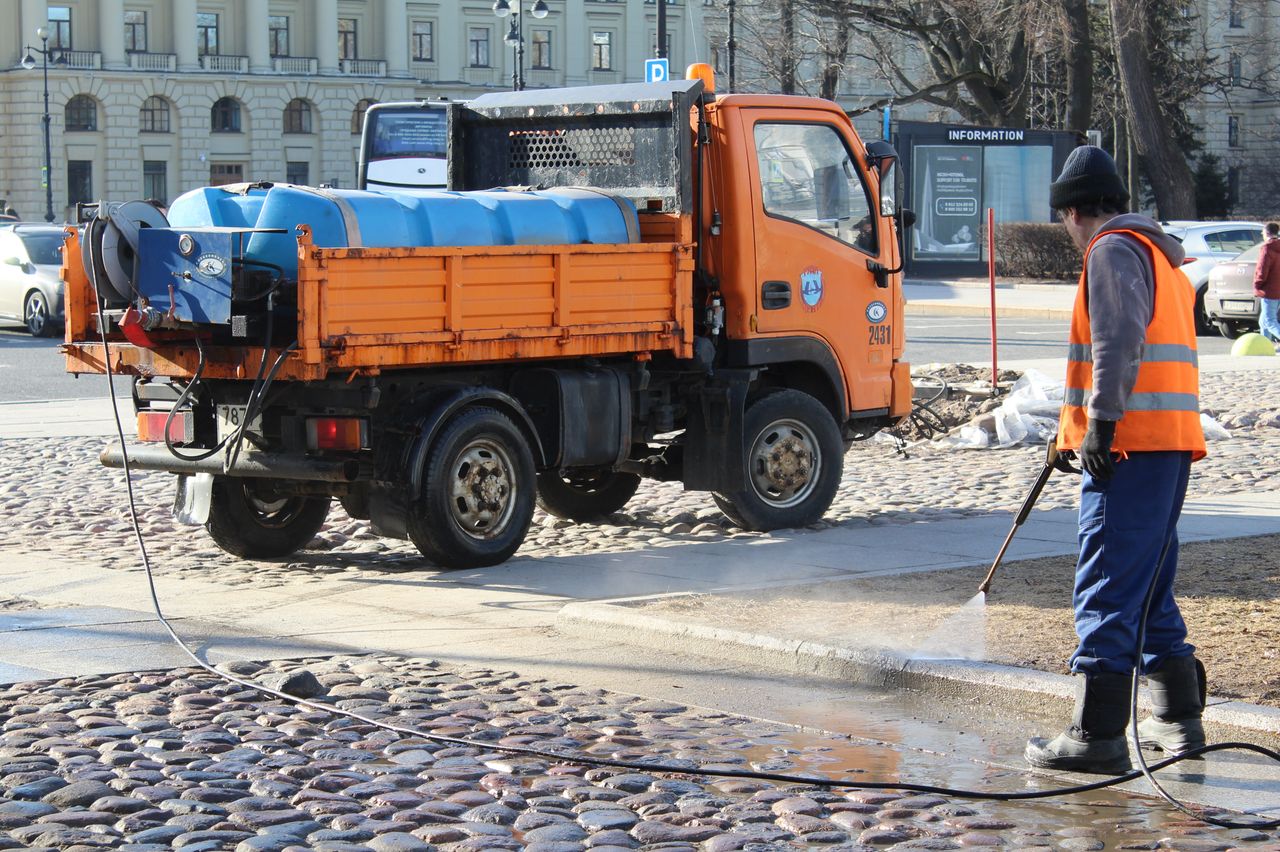services-01 A city worker cleans a cobblestone street with a hose and truck in an urban area.