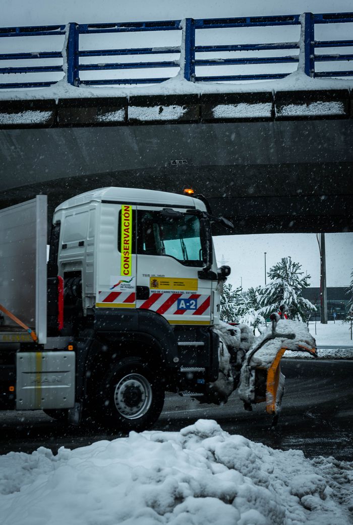 why-choose-us Snow plow truck clearing roads during a snowy winter day under a bridge in Guadalajara, Spain.