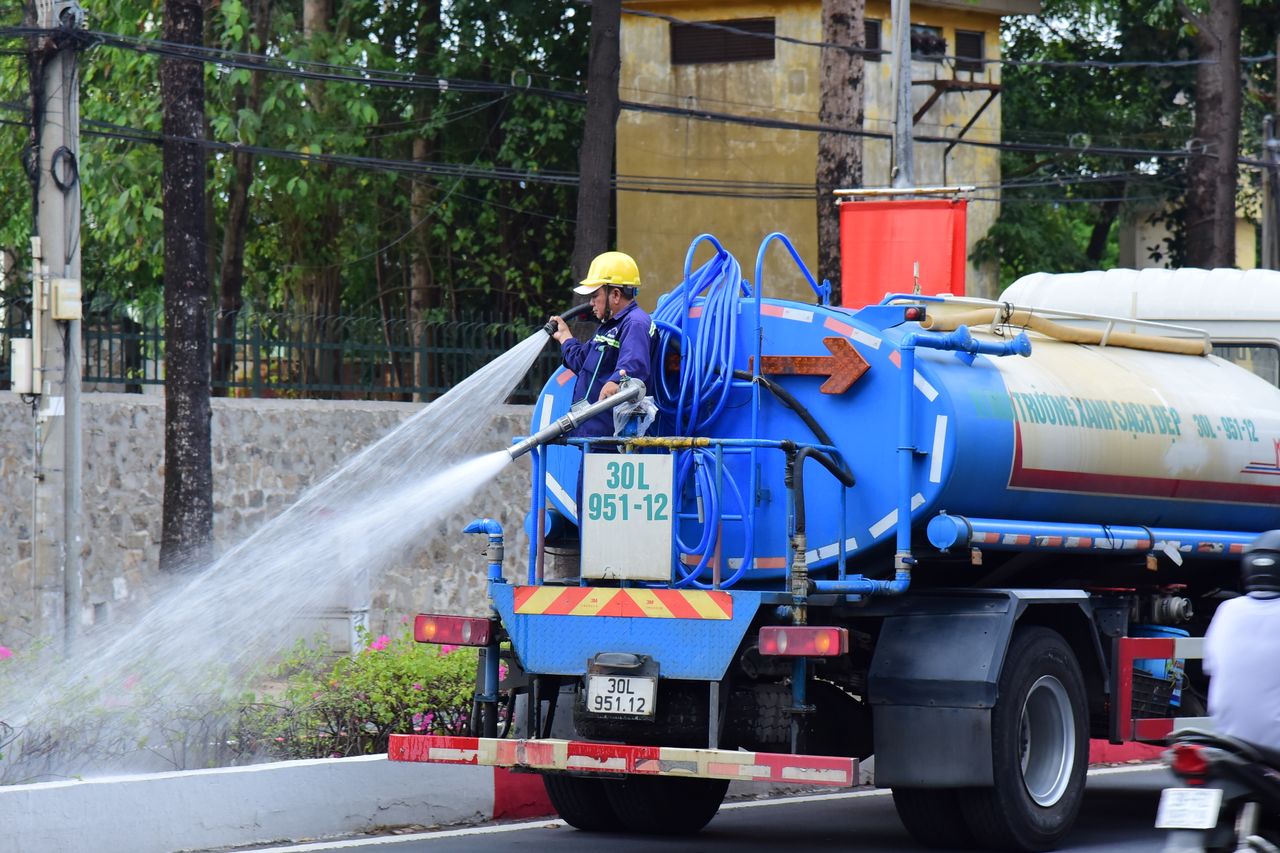 services-02 A worker operates a water truck in an urban setting, spraying water on the street.