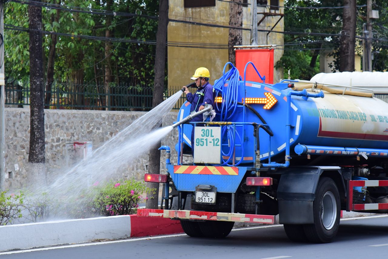 hero-img-02 Worker operates a water tanker spraying street plants for cleaning and maintenance.